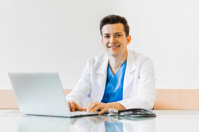 Photo of man in scrubs smiling in front of computer illustrates blog: "California Medical Legal Billing: What Is an EOR?