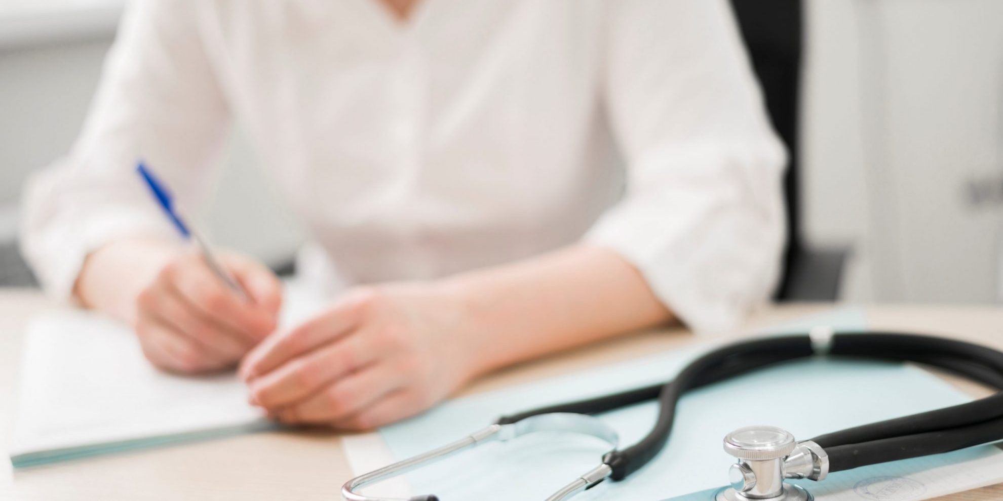 Practice-FP Woman in white shirt writing with stethoscope on the foreground.