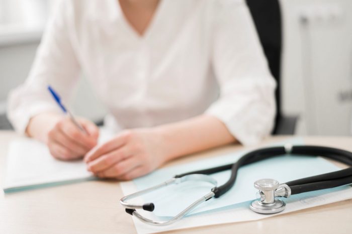 Practice-FP Woman in white shirt writing with stethoscope on the foreground.