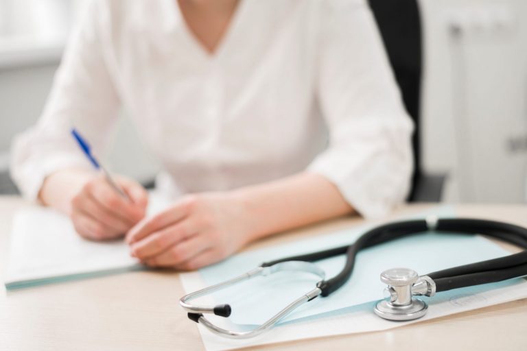 Woman in white shirt writing with stethoscope on the foreground.
