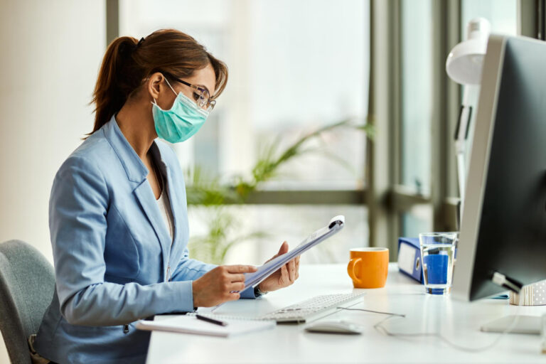 young-businesswoman-wearing-protective-face-mask-while-going-through-reports-working-office healthcare administrator analyzing revenue leakage healthcare data on computer